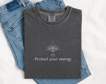 Puede incluir: Una camiseta gris oscuro con el texto "Protect your energy" y un gráfico blanco de un ojo, rayos de sol y una luna creciente. La camiseta está doblada sobre unos vaqueros azules.