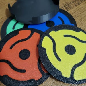 May include: A collection of colorful vinyl record adapters and a black record player spindle adapter. The adapters are in red, yellow, blue, and green, with a black center and a black border. The spindle adapter is black.