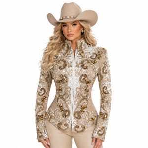 May include: A tan cowboy hat with a jeweled band sits atop a woman's head. She wears a tan jacket with intricate white and gold embroidery, a high collar, and a front zipper. The jacket is paired with matching tan pants.