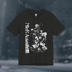 Peut inclure: T-shirt gris foncé avec un graphisme blanc du jeu vidéo Nier: Automata. Le design comprend le titre du jeu verticalement à gauche et une illustration détaillée de personnages au centre.