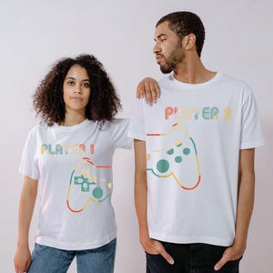 May include: Two white t-shirts featuring matching retro-style controller designs and the text "PLAYER 1" and "PLAYER 2" in pastel colours. The shirts are short-sleeved and worn by two people.