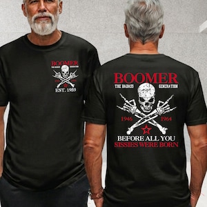 May include: A black t-shirt featuring a graphic print on the front and back. The design includes a skull, crossed skeletal hands, and red text reading BOOMER THE BADASS GENERATION, 1946, 1964, and BEFORE ALL YOU SISSIES WERE BORN.