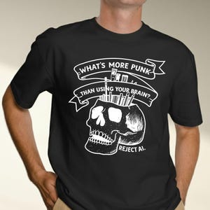 May include: A black t-shirt featuring a white graphic of a human skull with paintbrushes and pencils emerging from the top. Above the skull is a banner with the text "WHAT'S MORE PUNK THAN USING YOUR BRAIN?" and below the skull is the text "REJECT AI."