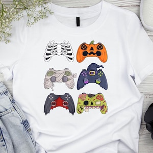 May include: White t-shirt featuring six Halloween-themed video game controller designs. The controllers include a skeleton, jack-o'-lantern, mummy, witch, bat, and zombie. The shirt is a casual, short-sleeved garment.