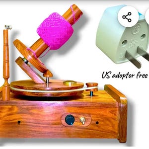 May include: A wooden yarn winder with a pink yarn ball. The winder has a wooden base, a rotating disc, and a vertical arm. A US adaptor is included.