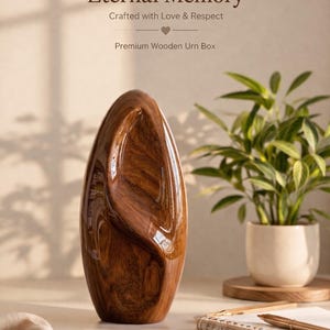Op de afbeelding: Een glanzende, druppelvormige houten urne met een rijke, donkerbruine afwerking. De tekst "Eternal Memory" en "Premium Wooden Urn Box" is zichtbaar. Een pot met een plant en briefpapier staan op de achtergrond.