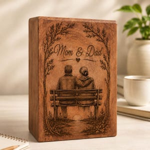Pu&ograve; includere: Un blocco di legno rettangolare con un'illustrazione incisa al laser di due persone sedute su una panchina sotto degli alberi. Il testo "Mom & Dad" &egrave; centrato sopra le figure con una piccola icona a forma di cuore. Il legno ha una finitura naturale.