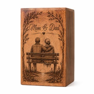 Pu&ograve; includere: Un'urna rettangolare in legno con un'illustrazione incisa al laser di due persone sedute su una panchina sotto degli alberi. Il testo Mom & Dad &egrave; centrato sopra le figure con un piccolo cuore. La scena include un sentiero e un sole.