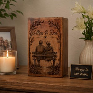 Pu&ograve; includere: Un'urna funeraria rettangolare in legno con un'illustrazione incisa al laser di due persone sedute su una panchina sotto degli alberi. Il testo "Mom & Dad" &egrave; inciso sopra e una piccola targa nera riporta "Always in our hearts."