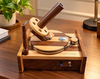 May include: A wooden yarn ball winder featuring a two-tone light and dark wood base, a rotating circular platform with a black drive belt, and a vertical spindle. The base includes a blue button and a metal toggle switch with the text Vür Andrea Lettmair.