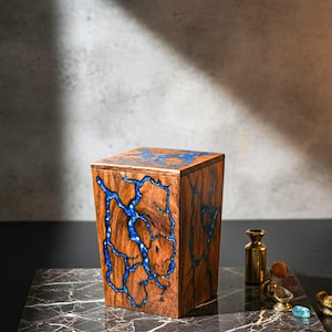 May include: A wooden box with a lid, featuring a unique design of blue resin veins running through the brown wood. The box is displayed on a marble-patterned surface, with small decorative items nearby. The box is approximately 6 inches tall.