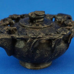 Könnte beinhalten: Eine dunkelgraue, aufwendig geschnitzte Teekanne mit Deckel und Ausguss. Die Teekanne weist florale und Rankenmuster auf und ist vor blauem Hintergrund platziert. Es ist ein dekorativer Artikel.