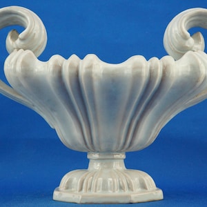 Könnte beinhalten: Hellblaue Keramikvase mit Sockel und zwei großen, geschwungenen Griffen. Die Vase hat ein geriffeltes Design und ist vor einem blauen Hintergrund platziert. Die Vase ist etwa 30 cm hoch.