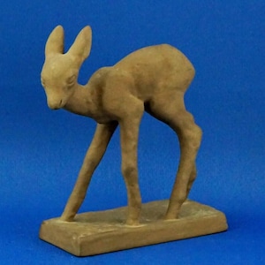 May include: A tan-colored ceramic sculpture of a young deer, standing on a rectangular base. The deer has large ears and slender legs, with a slightly curved posture. The sculpture is set against a blue background.