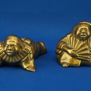 以下が含まれることがあります： 青い背景に2つの金色の置物。1つは横たわり、もう1つは座って扇子を持っています。置物は詳細な顔の特徴があり、装飾品である可能性があります。