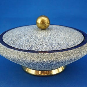 Pode incluir: Uma tigela decorativa com tampa, exterior salpicado em azul e cinza, acentuada por uma faixa azul escura e uma base dourada. A tampa é encimada por um pequeno botão redondo dourado. A tigela está sobre um fundo azul liso.