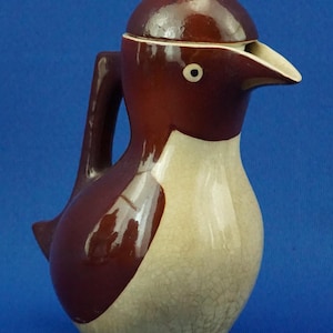 May include: A ceramic pitcher in the shape of a penguin, featuring a dark brown head and back, a cream-colored belly, and a white beak. The pitcher has a handle and a lid, and is set on three small feet. The design is whimsical and decorative.