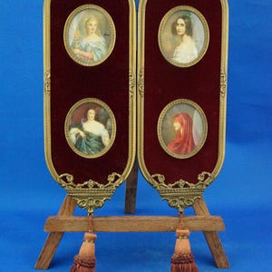 May include: A pair of antique portrait frames with oval miniature paintings. Each frame has a burgundy velvet backing, ornate gold-colored metal trim, and a decorative tassel. The frames are displayed on a wooden easel.