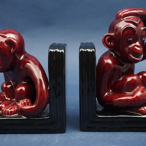 Puede incluir: Par de sujetalibros de mono de cerámica. Cada sujetalibros presenta una figura de mono brillante de color granate sobre una base negra. Un mono está sentado, el otro apoya la cabeza en su mano. Decoración del hogar.