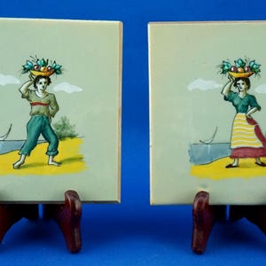 Peut inclure: Deux carreaux de céramique carrés avec des scènes peintes de personnages portant des paniers de fruits sur la tête. Les personnages sont représentés près d'une plage avec un voilier et des arbres. Les carreaux sont exposés sur des présentoirs en bois.