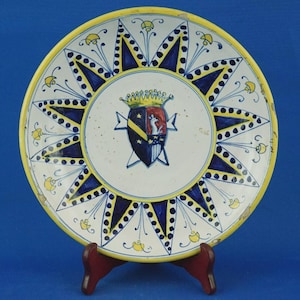 Op de afbeelding: Een decoratieve keramische plaat met een gele rand en een centraal embleem. De plaat heeft een sterrenpatroon in blauw en geel, rondom een wapen met een kroon en schild. De plaat wordt gepresenteerd op een houten standaard.