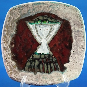 May include: Square ceramic plate with a stylized figure design. The plate features a central image of a figure with a white body, red background, and a textured, speckled border. The figure has a green and black headpiece.