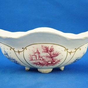 May include: White ceramic bowl with a scalloped rim and three small feet. The bowl is decorated with red, blue, and gold landscape scenes. The rim has a gold trim. The bowl is on a blue background.