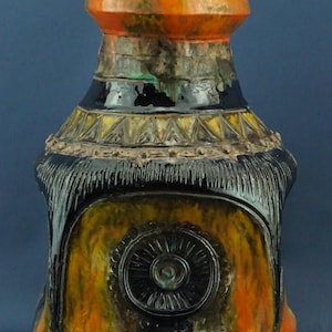 May include: A tall, decorative ceramic vase with a unique, textured design. The vase features a color palette of orange, black, and yellow. The top is orange, transitioning to black and yellow with intricate patterns and a central circular design.