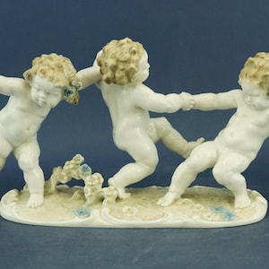 Puede incluir: Figura de porcelana que representa a tres querubines en una pose juguetona, tomados de la mano y corriendo. Las figuras tienen rasgos faciales detallados y cabello rizado. La base es blanquecina con sutiles detalles azules.