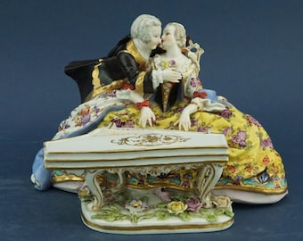 May include: A porcelain figurine of a couple in 18th-century dress, seated near a decorative piano. The woman wears a yellow floral dress, the man a black coat. The piano is white with gold detailing. The figurine is a decorative item.