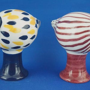 以下が含まれることがあります： 鳥の形をした2つのセラミック製の塩コショウ入れ。1つは白地に黄色とネイビーブルーのアクセント、もう1つは白地に赤いストライプ。どちらもネイビーブルーのキャップと黄色のくちばしが付いています。シェーカーは濃い青と赤のベースにあります。
