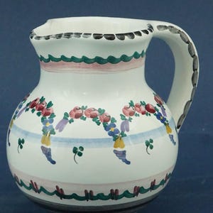 May include: White ceramic jug with a handle, decorated with floral garlands in pink, red, and blue. The jug has green and pink wavy bands, and black accents. A decorative piece for serving beverages.