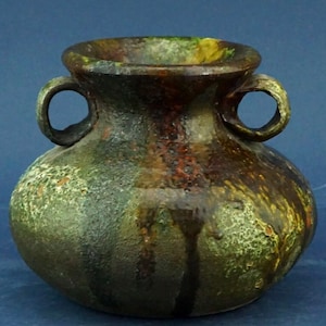 Może przedstawiać: Mały, ręcznie robiony ceramiczny wazon z dwoma uchwytami w kształcie pętli. Wazon ma teksturowaną powierzchnię z mieszanką zielonych, brązowych i żółtych odcieni. Projekt obejmuje ciemny, spływający efekt.