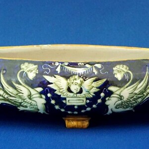 May include: A long, oval ceramic planter with a dark blue exterior, adorned with white, green, and yellow floral and cherub designs. The planter has small feet and handles on each end. The interior is a light cream color.