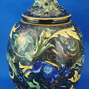 May include: A decorative ceramic jar with a lid, featuring a dark blue background and colorful hand-painted details. The jar showcases a running blue hare amidst foliage, with yellow, green, and brown accents. The lid is topped with a black finial.