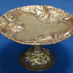 May include: A vintage brass pedestal dish with a round, floral-embossed top and a matching base. The dish has a tarnished gold finish, with intricate leaf and flower details around the edges and base. The pedestal dish is set against a blue background.