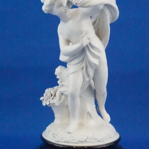 May include: A white porcelain figurine depicting a classical figure draped in fabric, standing on a textured base. The figure is posed with a flowing cloth, and a small figure is at the base. The base is black with gold trim.