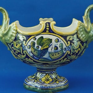 May include: A ceramic bowl with a pedestal base and two loop handles. The bowl is decorated with blue, yellow, and green floral and figure designs. The text "EL VIA" is visible on the front.