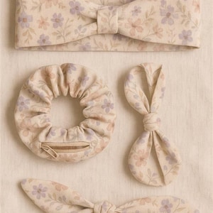 Puede incluir: Un conjunto de accesorios para el cabello con estampado floral en crema, rosa y lila. El conjunto incluye una diadema, una goma para el pelo con cremallera y dos lazos para el pelo. La tela tiene un delicado estampado floral.