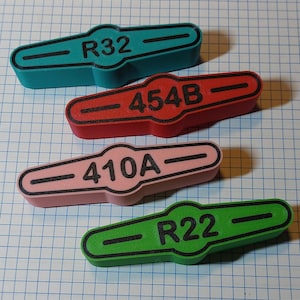 Peut inclure: Quatre étiquettes rectangulaires colorées pour vannes de CVC. Les étiquettes sont turquoise, rouge, rose et verte. Chaque étiquette porte le texte noir : "R32", "454B", "410A" et "R22". Les étiquettes sont sur un fond de grille bleu.