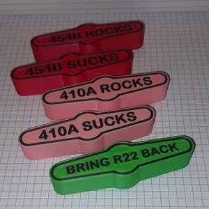 Könnte beinhalten: Eine Sammlung farbenfroher, rechteckiger 3D-gedruckter Objekte mit abgerundeten Enden. Die Objekte sind rot, rosa und grün und tragen jeweils schwarzen Text. Der Text enthält Sätze wie "454B ROCKS", "454B SUCKS", "410A ROCKS", "410A SUCKS" und "BRING R22 BACK".