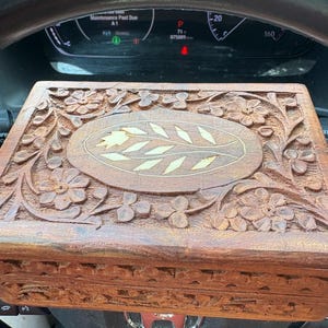 May include: A brown wooden box with intricate floral carvings. The box features a central oval design with a light-colored leaf pattern. The box is rectangular and appears to be handmade, showcasing detailed craftsmanship.