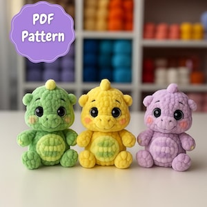 May include: Three crocheted dinosaur plushies in green, yellow, and purple. Each has large black eyes, rosy cheeks, and a smiling expression. The plushies are sitting on a white surface. A purple sign above reads "PDF Pattern".