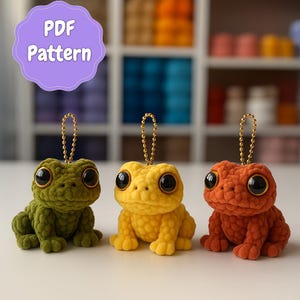 Könnte beinhalten: Drei gehäkelte Frosch-Schlüsselanhänger in Grün, Gelb und Orange. Jeder Frosch hat große schwarze Augen und eine goldene Kette. Das Bild enthält ein lila Schild mit der Aufschrift "PDF Pattern".