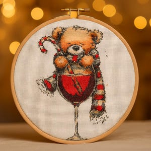 May include: Cross-stitch embroidery in a wooden hoop features a teddy bear holding a candy cane and wearing a scarf, inside a wine glass. The bear is light brown, the scarf is red and white, and the wine is red.