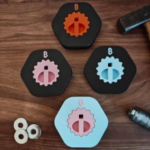Puede incluir: Un conjunto de coloridos tapones hexagonales para botellas. Cada tapón presenta un diseño único con un inserto central de color y un anillo exterior decorativo. Los tapones son negros, naranjas, rosas y azules, con un pequeño logotipo "B".