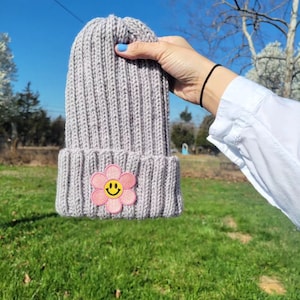 May include: A gray knitted beanie with a pink flower patch featuring a yellow smiley face.