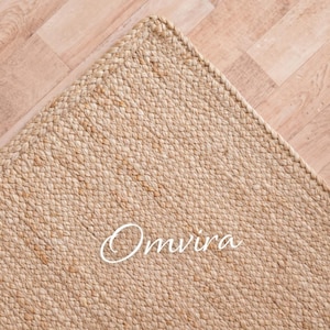 May include: A corner of a beige jute rug with a braided edge. The rug is on a light brown wooden floor. The word "Omvira" is written in cursive on the rug.