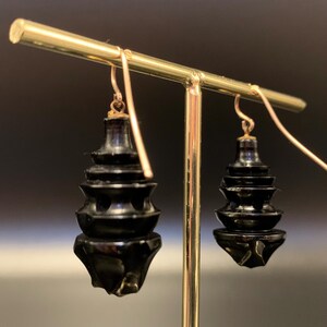 May include: A pair of black, tiered drop earrings with gold-colored ear wires. The earrings have a pagoda-like design with multiple levels and a faceted bottom. The earrings are displayed on a gold-colored jewelry stand against a dark background.