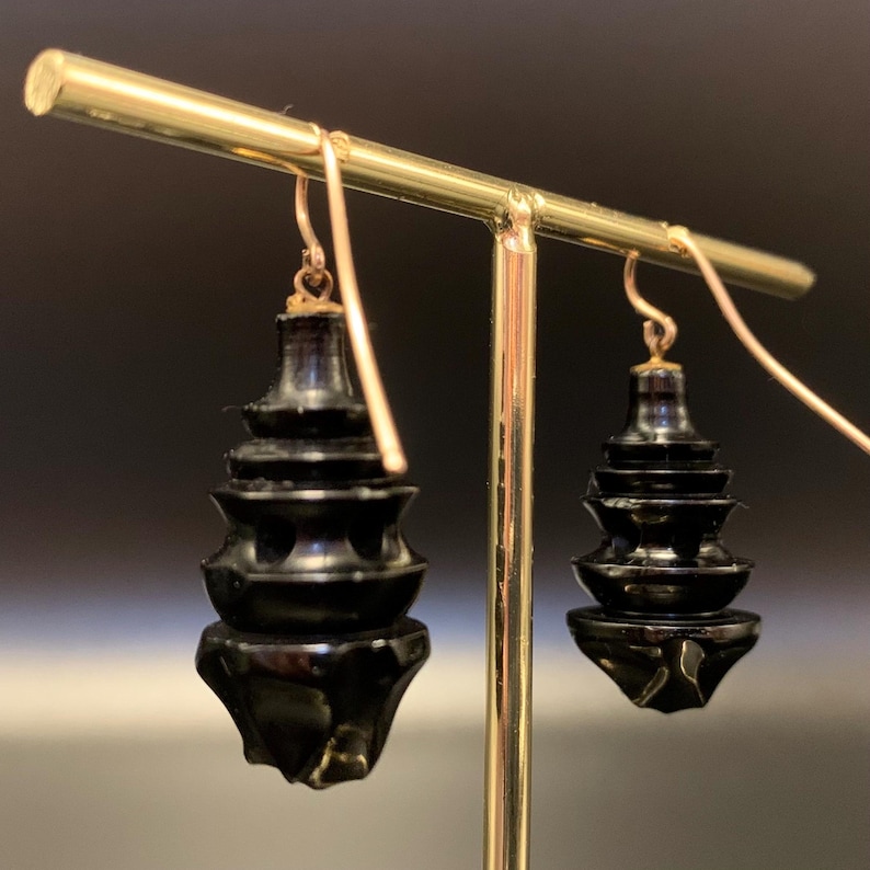 May include: A pair of black, tiered drop earrings with gold-colored ear wires. The earrings have a pagoda-like design with multiple levels and a faceted bottom. The earrings are displayed on a gold-colored jewelry stand against a dark background.